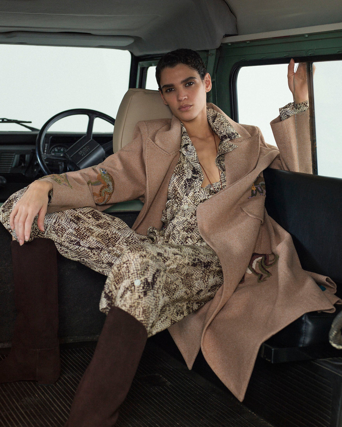 A person sits in the back of a vehicle wearing a patterned jumpsuit, a long tan coat with embroidered details, and knee-high brown boots, with one arm resting on the seat.