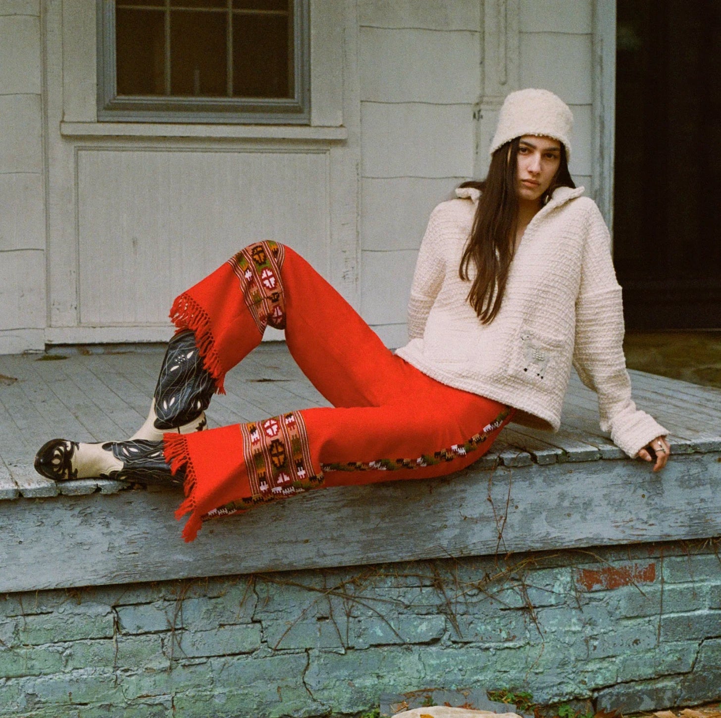 A person in a cozy wool sweater and hat sits on a porch. They wear vibrant red pants with decorative patterns and tasseled fringes, paired with black and white heeled boots. The porch has a weathered, rustic look.