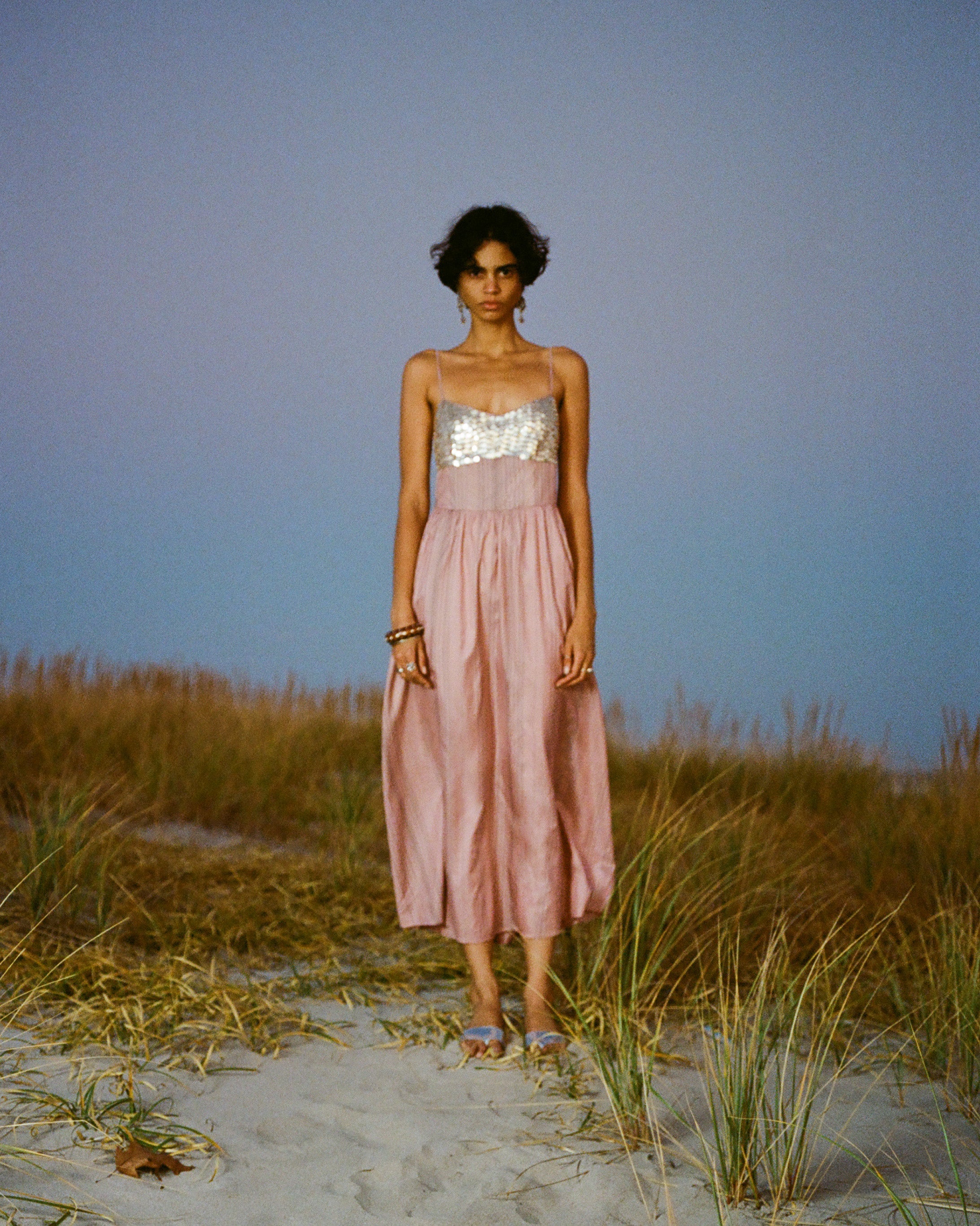 A person stands on sand surrounded by grass at dusk. They wear a pink dress with a silver, sequined top.