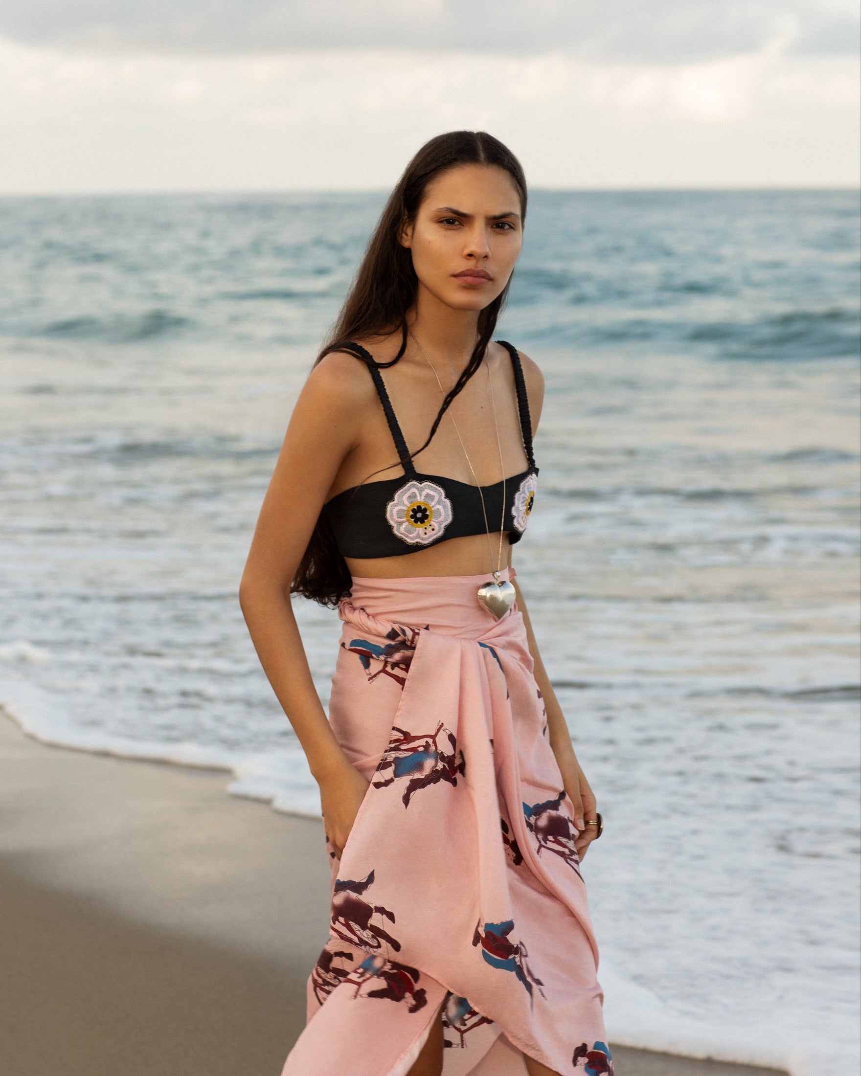 A woman in a floral bralette and a pink skirt with horse prints stands on a beach near the shoreline, with waves and cloudy sky in the background.