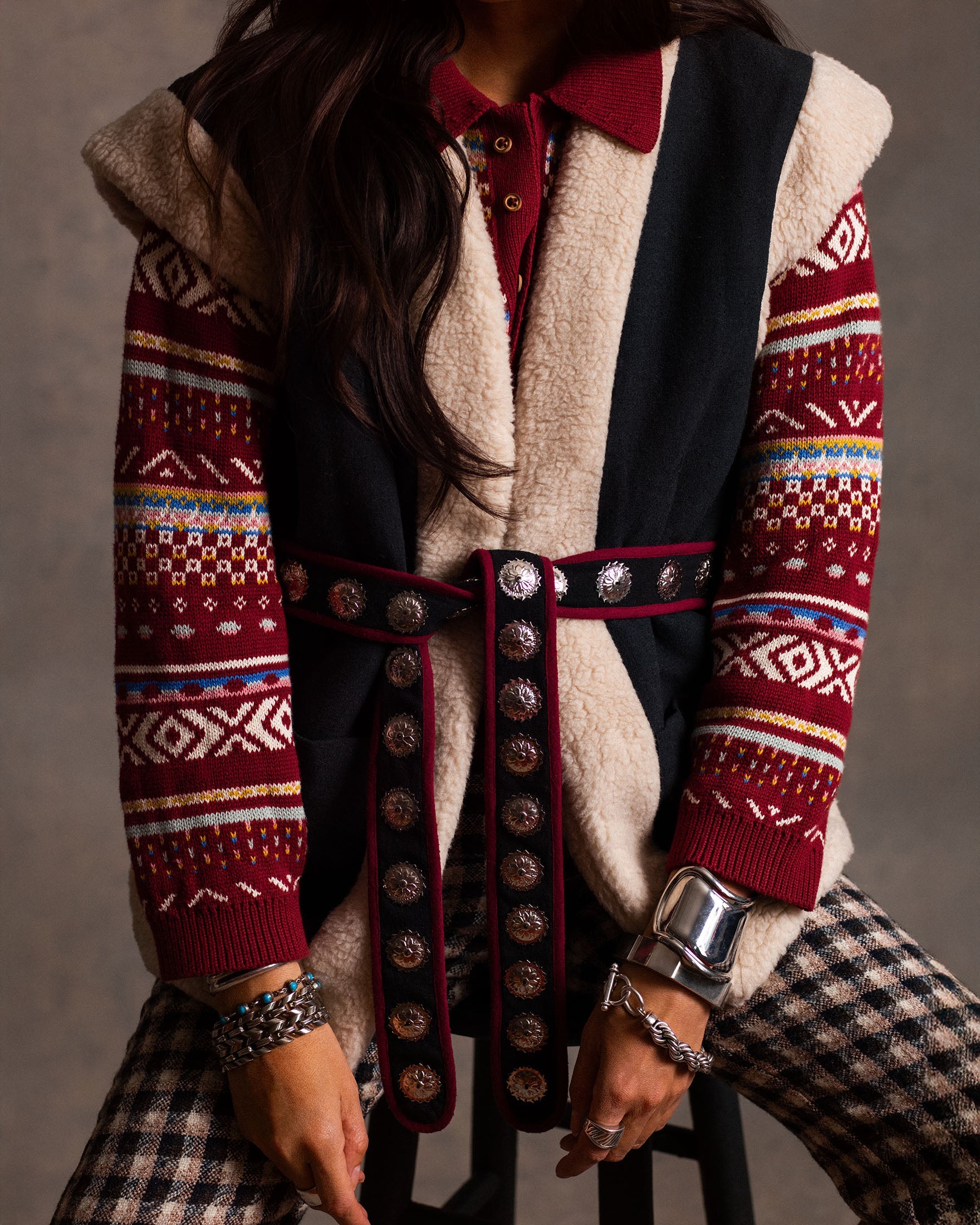 A person wearing a patterned red sweater, a sleeveless shearling vest, a wide decorative belt, plaid pants, and multiple silver bracelets sits on a stool.