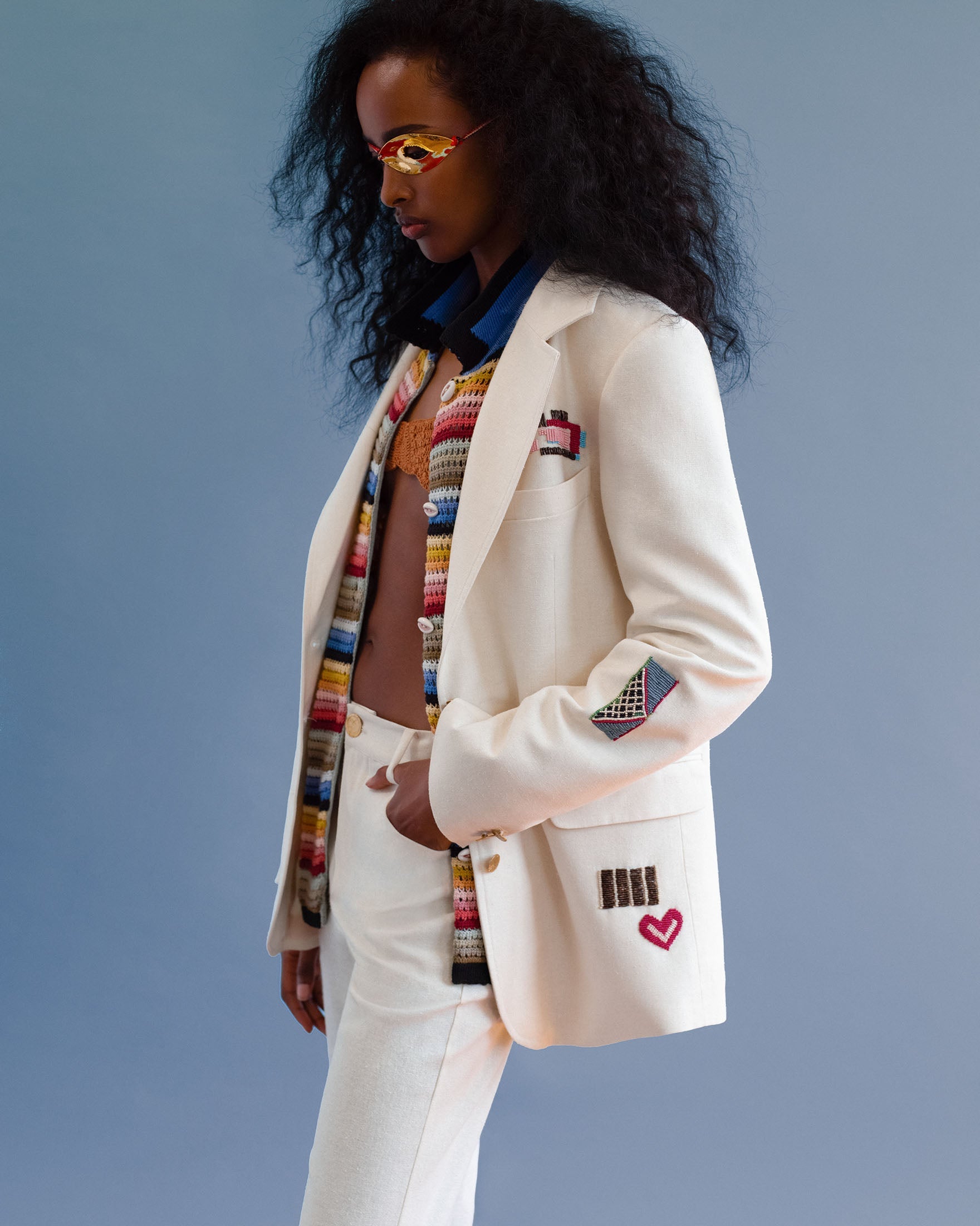 A person wearing a cream-colored suit with embroidered patches, a striped crochet top underneath, and yellow sunglasses, standing against a blue background.