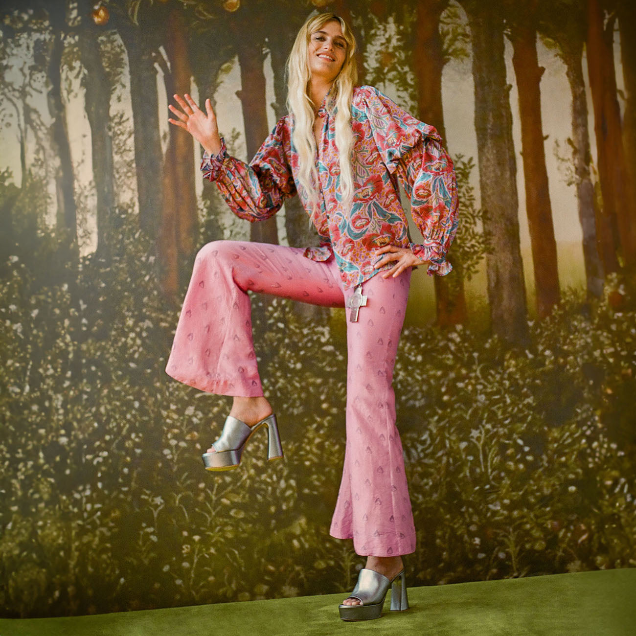 A woman in a paisley blouse and pink flared pants poses with one knee raised, wearing platform sandals, against a forest-themed backdrop.