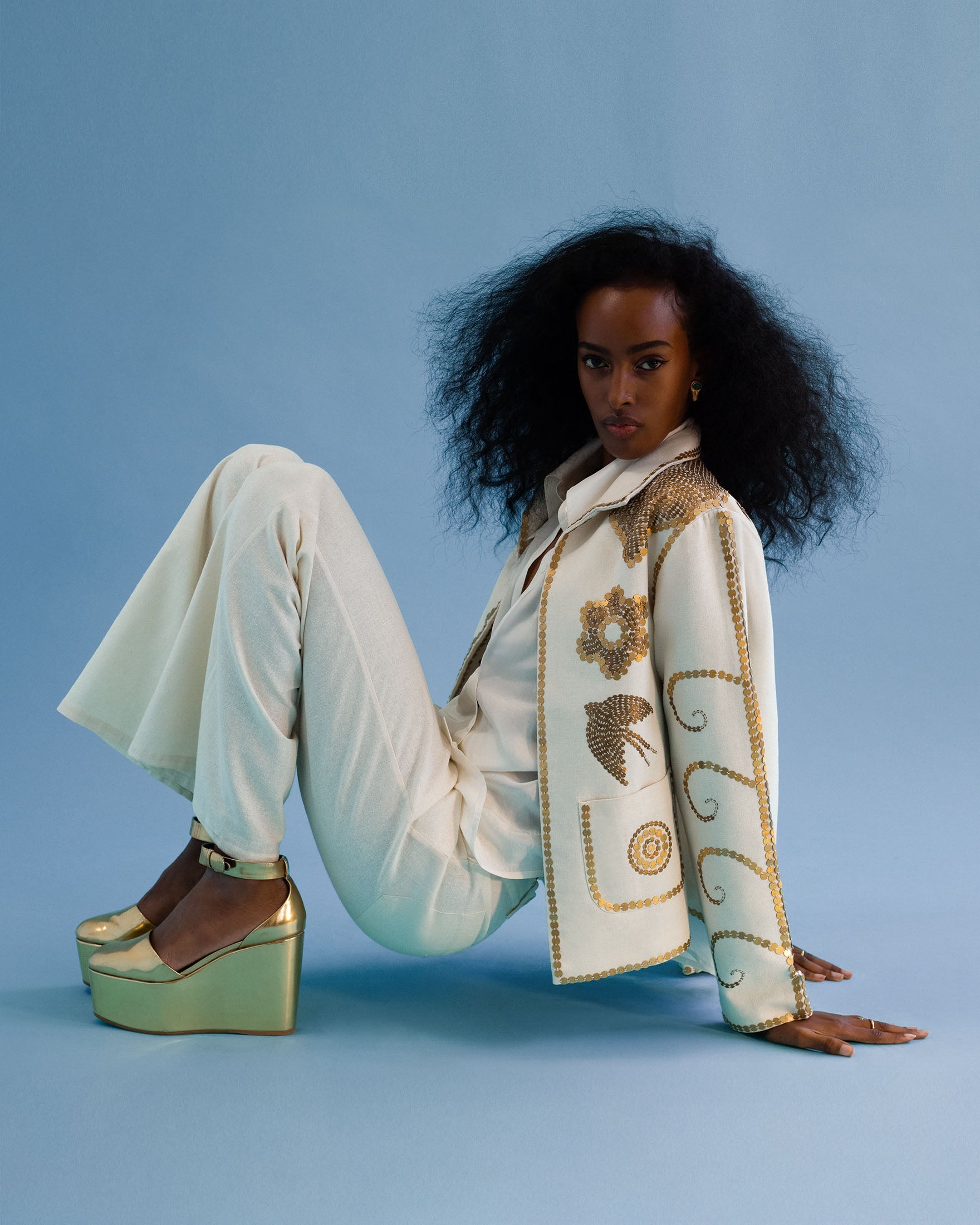 A woman in a paisley blouse and pink flared pants poses with one knee raised, wearing platform sandals, against a forest-themed backdrop.