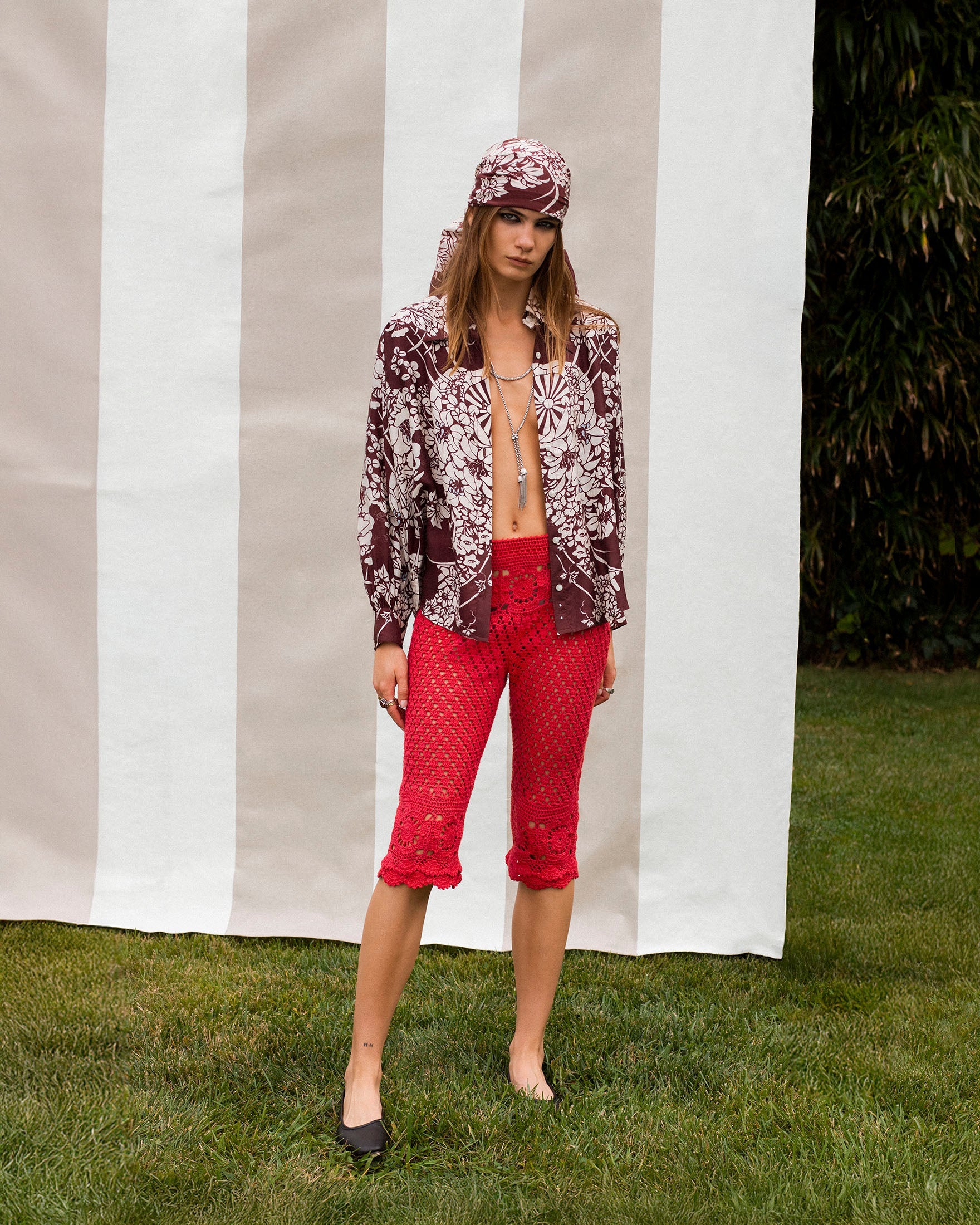 A person stands on grass in front of a striped backdrop, wearing a patterned bandana, matching shirt, red lace cropped pants, and black flats.
