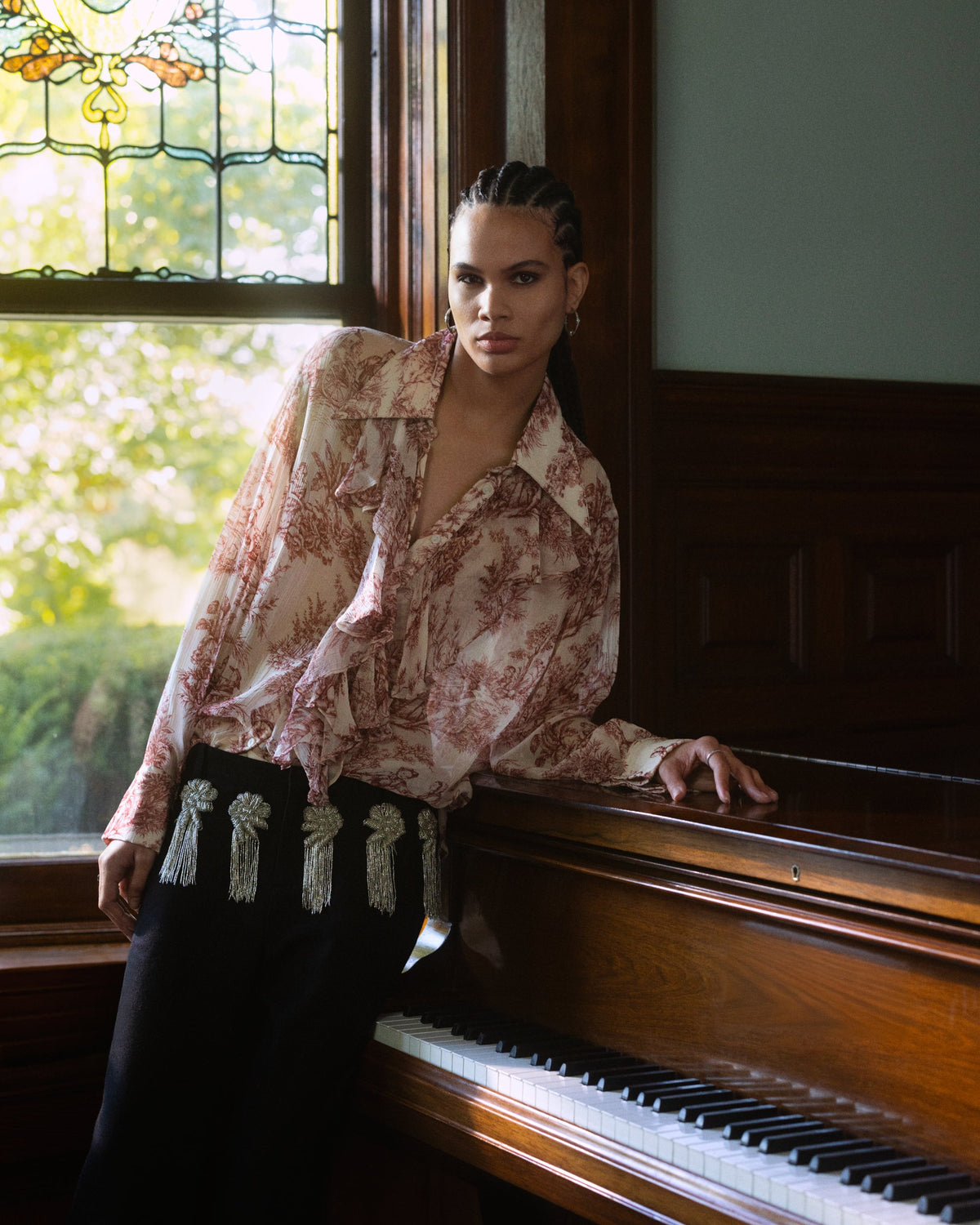 A person with braided hair wears a ruffled, patterned blouse and black trousers with silver fringe detailing, leaning against a wooden piano by a window.