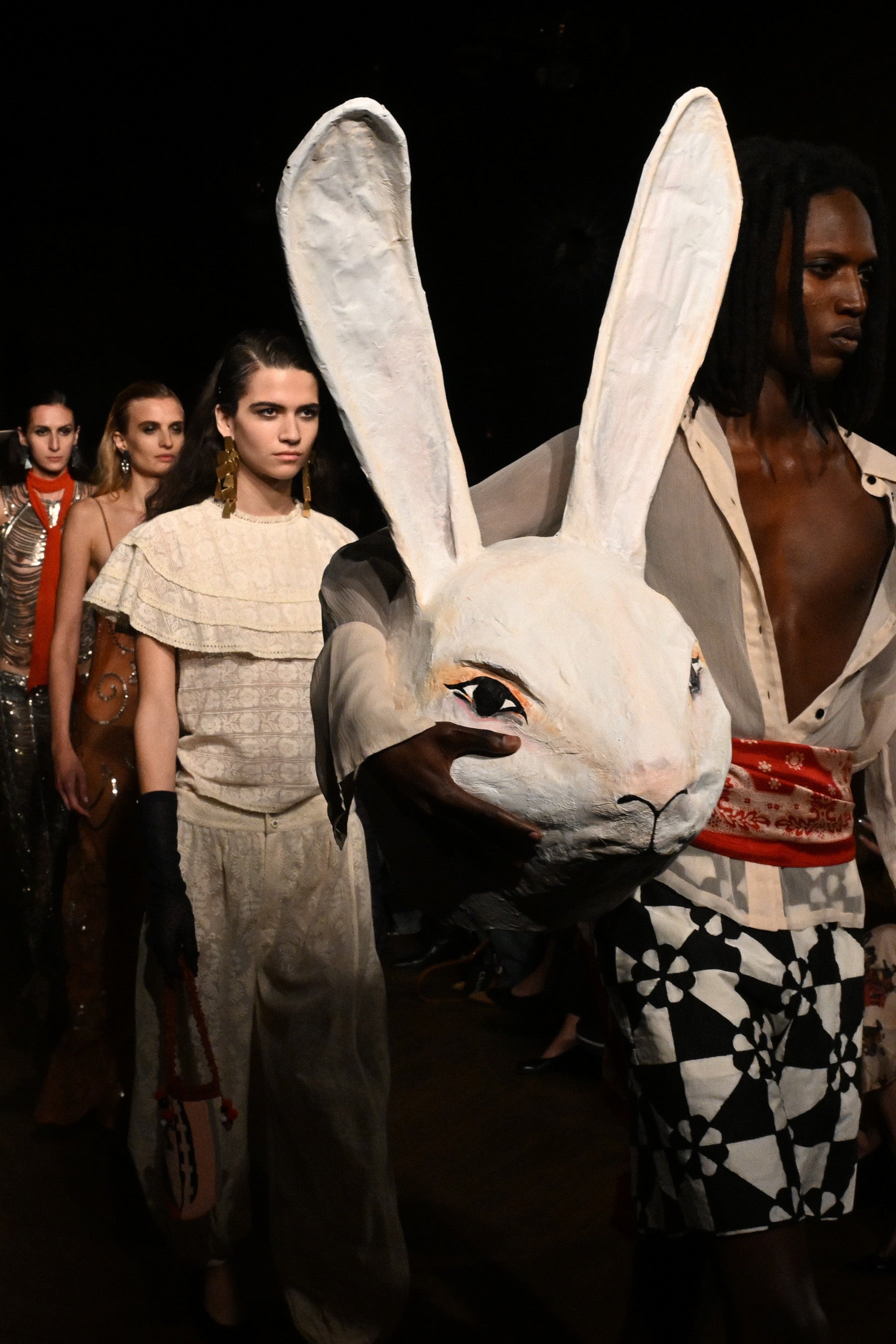Models walk down a runway in eclectic outfits, with one person carrying a large papier-mâché rabbit head.