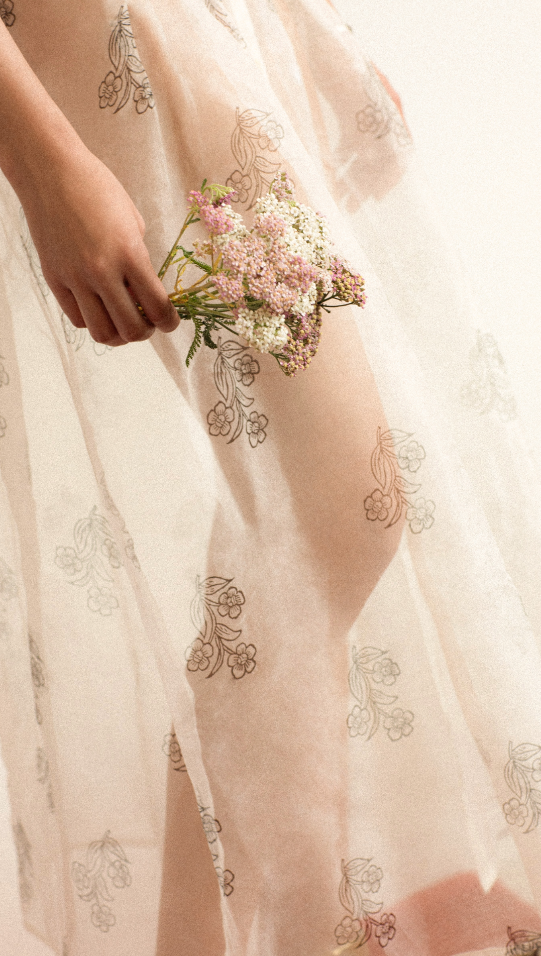 A person in a sleeveless, ruffled dress with a wide belt stands against a plain background, raising one arm to partially cover their face.
