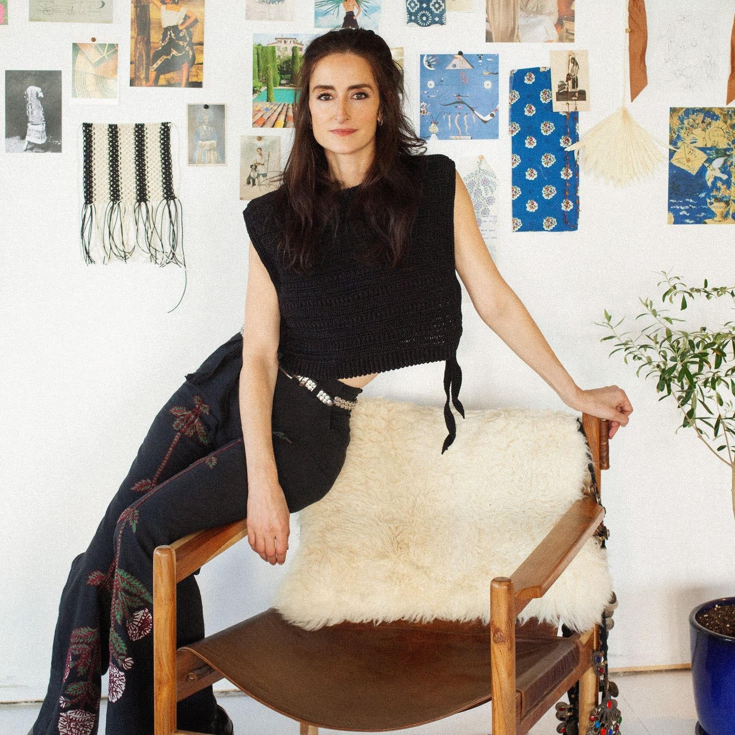 A woman in a black sleeveless top and patterned pants sits on the arm of a chair with a fur throw, in front of a wall covered with various art pieces and swatches.