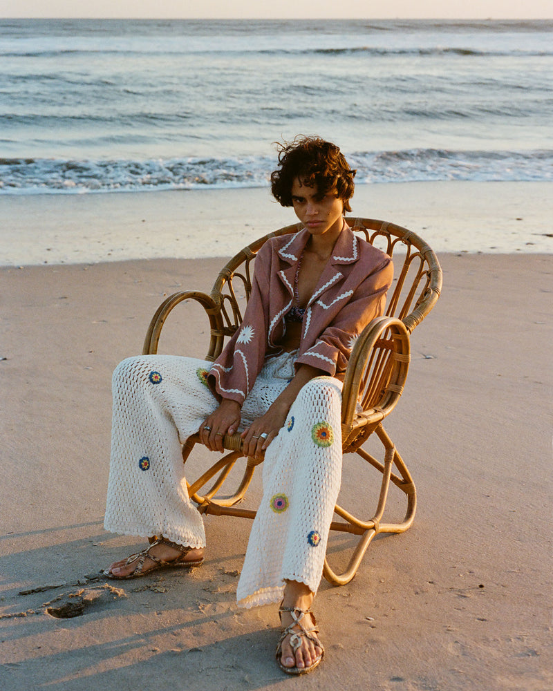 A person with short hair sits in a wicker chair on a sandy beach at sunset. They wear a patterned, long-sleeved top and white crochet pants with colorful flower accents, along with sandals. The ocean waves are in the background.