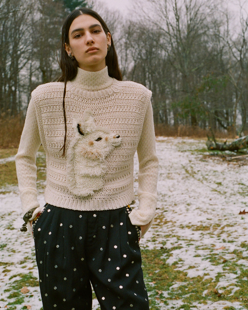 A person stands outdoors in a snowy, wooded area. They are wearing a cream knit sweater featuring a 3D animal head design on the front and dark pants with a dotted pattern. Their expression is neutral, and their hands are in their pockets.