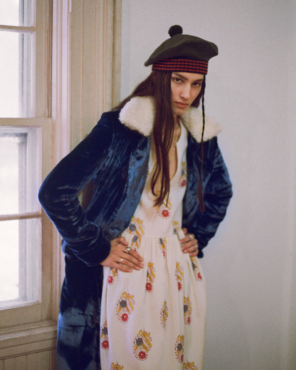 A person stands indoors wearing a long black coat with a white fur collar over a floral-patterned dress. They have long hair and wear a green beret. Their hands are on their hips, and they are looking towards the camera.