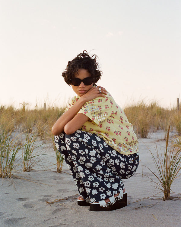 A person in sunglasses crouches on a sandy beach, wearing a yellow floral top and black floral pants. The background features tall grasses under a clear sky.