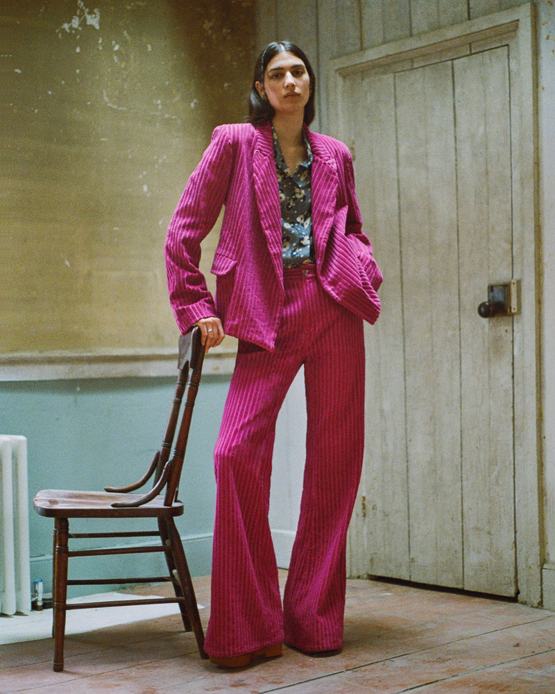 A person wearing a vibrant pink corduroy suit and patterned shirt stands confidently in a vintage-style room with peeling paint and a wooden chair. They have long dark hair and are posing with one hand in their pocket.