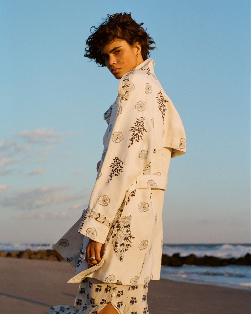 A person with short, curly hair stands on a beach at sunset, wearing a light-colored coat with a floral pattern. The beach has gentle waves in the background under a clear sky.