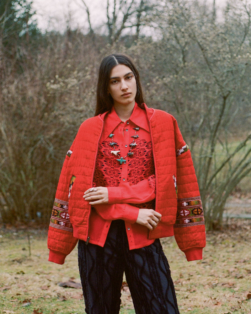 A person stands outdoors wearing a red padded jacket over a red shirt with a detailed pattern. They are also wearing black pants. The background is a natural setting with bushes and trees.