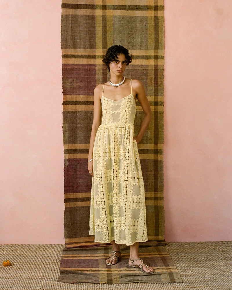 A person wearing a cream-colored, sleeveless crochet dress stands on a woven mat against a striped textile backdrop. They have short curly hair and are accessorized with sandals and necklaces. The setting has a soft, warm tone.