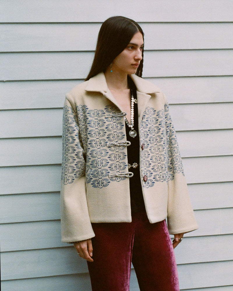 A woman with long dark hair stands against a light gray wall. She wears a cream jacket with blue floral patterns, a black top with a pearl necklace, and dark red velvet pants.