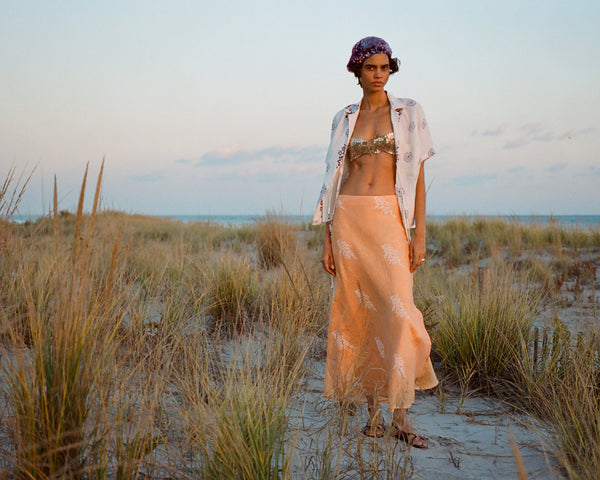 A person stands on a sandy beach surrounded by tall grass. They are wearing a patterned headscarf, open shirt, decorative top, and a flowing peach-colored skirt. The sky is clear with a subtle sunset hue.