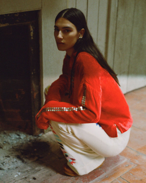 A person with long dark hair crouches by a fireplace, wearing a bright red top with sequined stripes on the sleeves and patterned white pants. The setting appears to be a dimly lit room with wooden paneling and brick flooring.