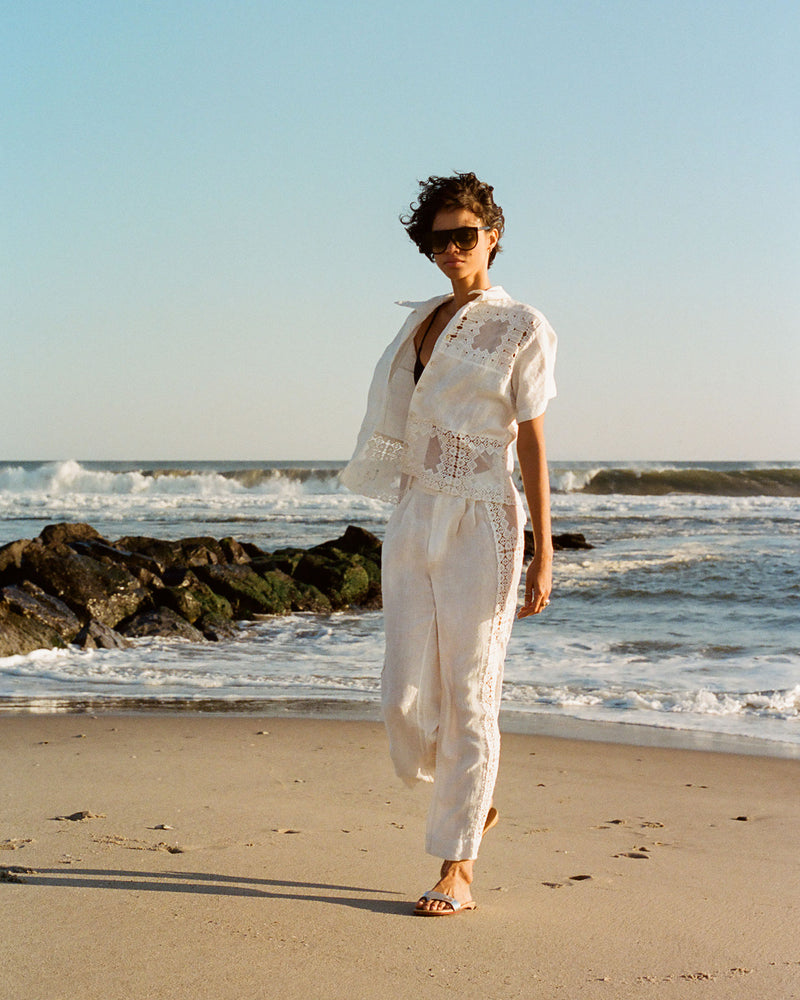 A person in a white outfit walks barefoot along a sandy beach with ocean waves in the background under a clear blue sky. They have short, curly hair and are wearing sunglasses.