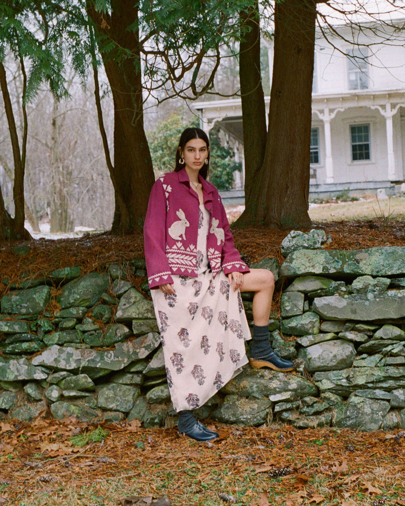 A person stands in front of a stone wall surrounded by trees, wearing a patterned maroon jacket and a long beige skirt with designs. They are outdoors with a house partially visible in the background. The ground is covered in leaves.