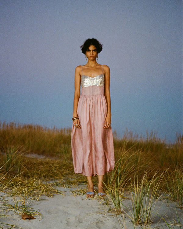 A woman in a pink dress with a silver sequin top stands on a sandy path surrounded by tall grasses under a twilight sky.