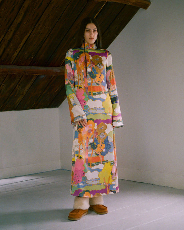 A person stands indoors wearing a long, colorful, patterned dress featuring abstract and floral designs. The ceiling has exposed wooden beams, and the person is wearing brown shoes with a light-colored sole.