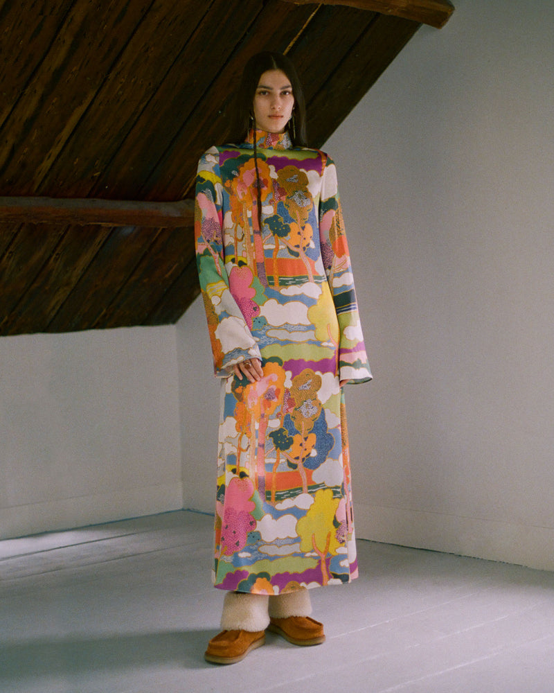 A person stands indoors wearing a long, colorful, patterned dress featuring abstract and floral designs. The ceiling has exposed wooden beams, and the person is wearing brown shoes with a light-colored sole.