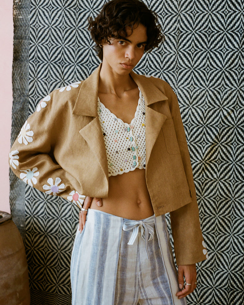 A person stands confidently wearing a brown jacket with white flower appliqués, a crocheted white top, and striped pants. The background features a geometric black and white pattern.