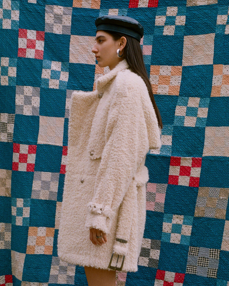 A person in a woolly, oversized coat and black cap stands in profile against a patchwork quilt background with blue, beige, and red squares. They have long hair and are wearing hoop earrings.