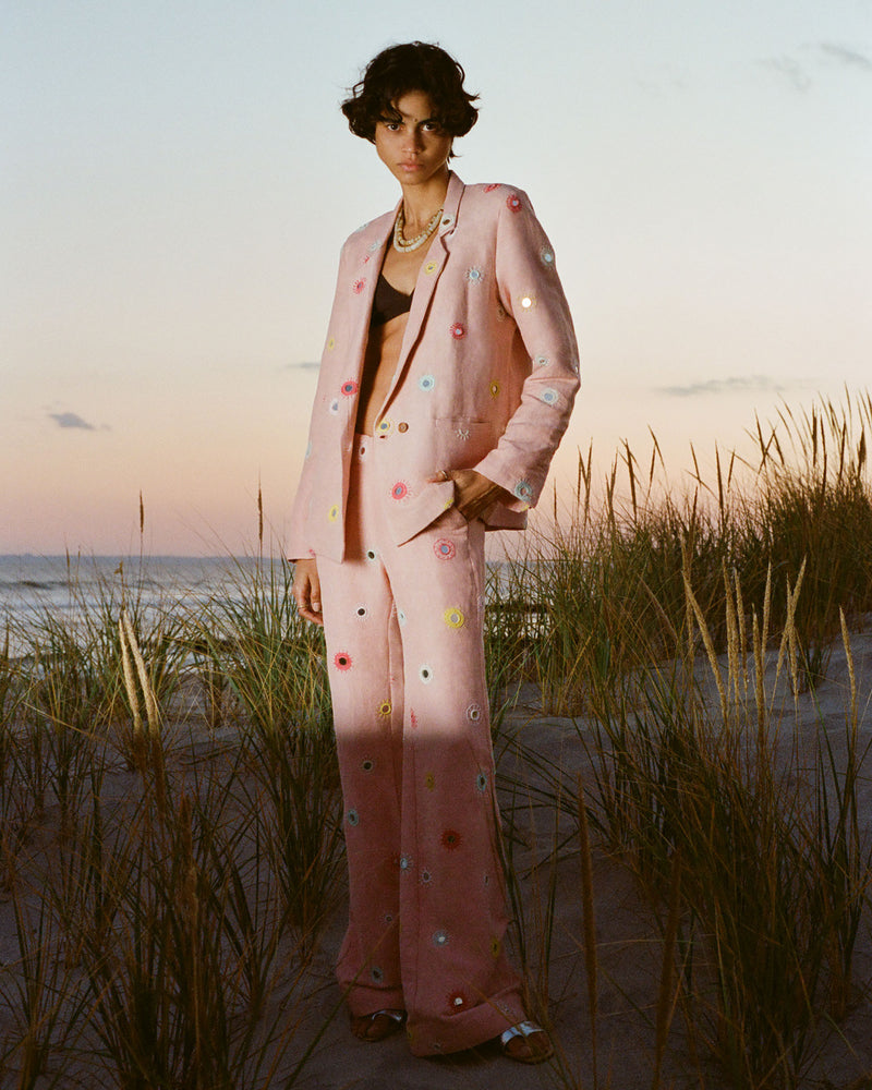 A person stands on a sandy beach at sunset, wearing a pink suit adorned with colorful circular embellishments. Tall grasses surround them, with the ocean in the background.