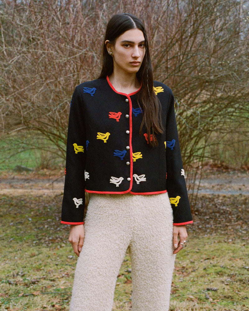 A woman stands outdoors in a wooded area, wearing a black jacket with colorful bird patterns and red trim, paired with cream textured pants. The background features bare trees and grass.