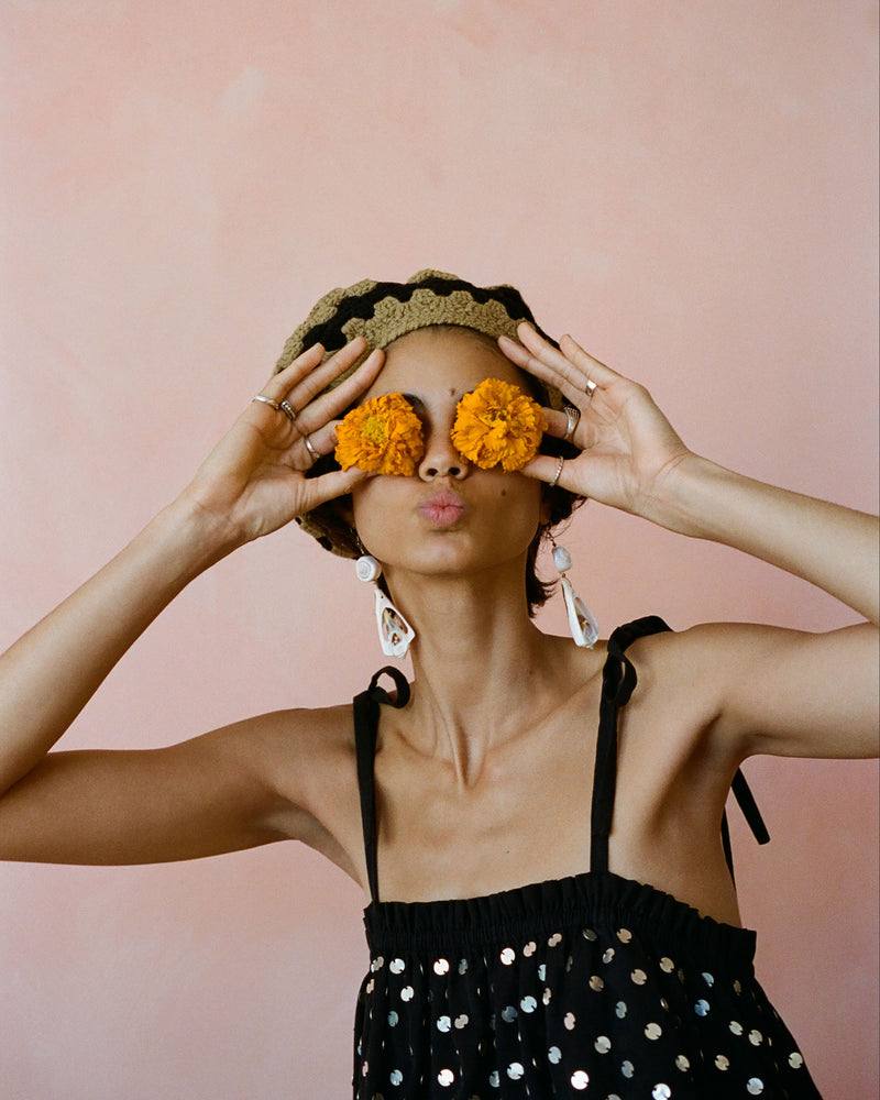 A person playfully holds two yellow flowers over their eyes, puckering their lips. They wear a black, polka-dotted dress with tie straps and a knitted hat. The background is a soft pink color.