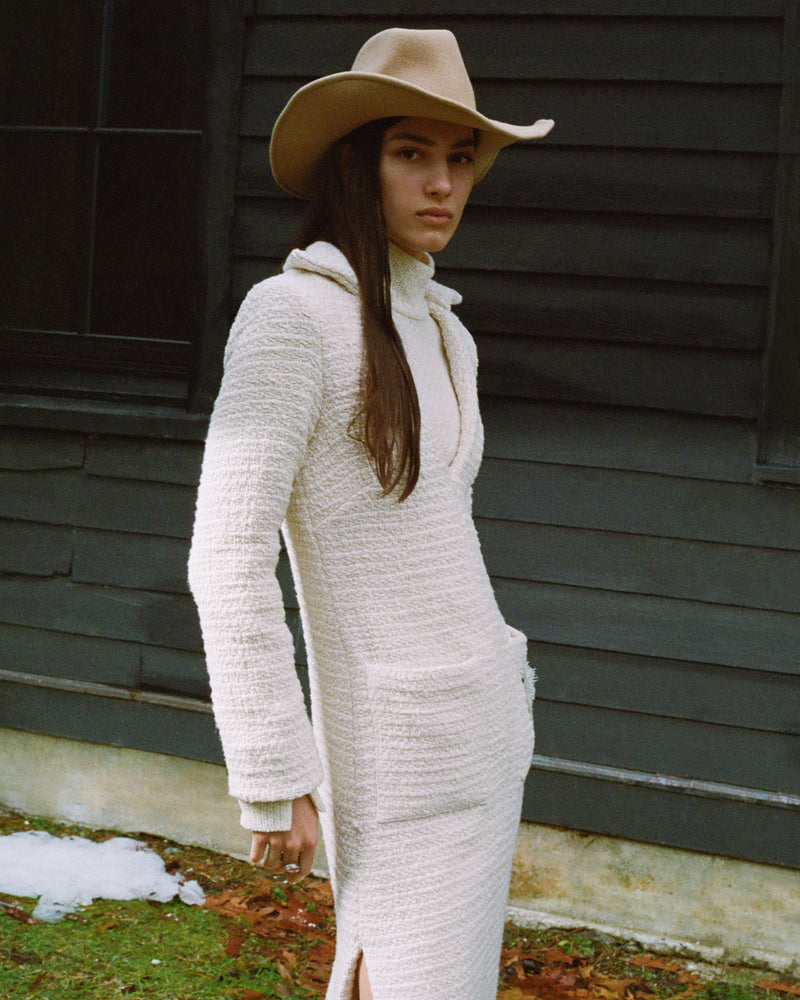 A person with long dark hair wearing a beige cowboy hat and a textured long-sleeve dress stands in front of a dark wooden building. Snow and fallen leaves are visible on the ground.