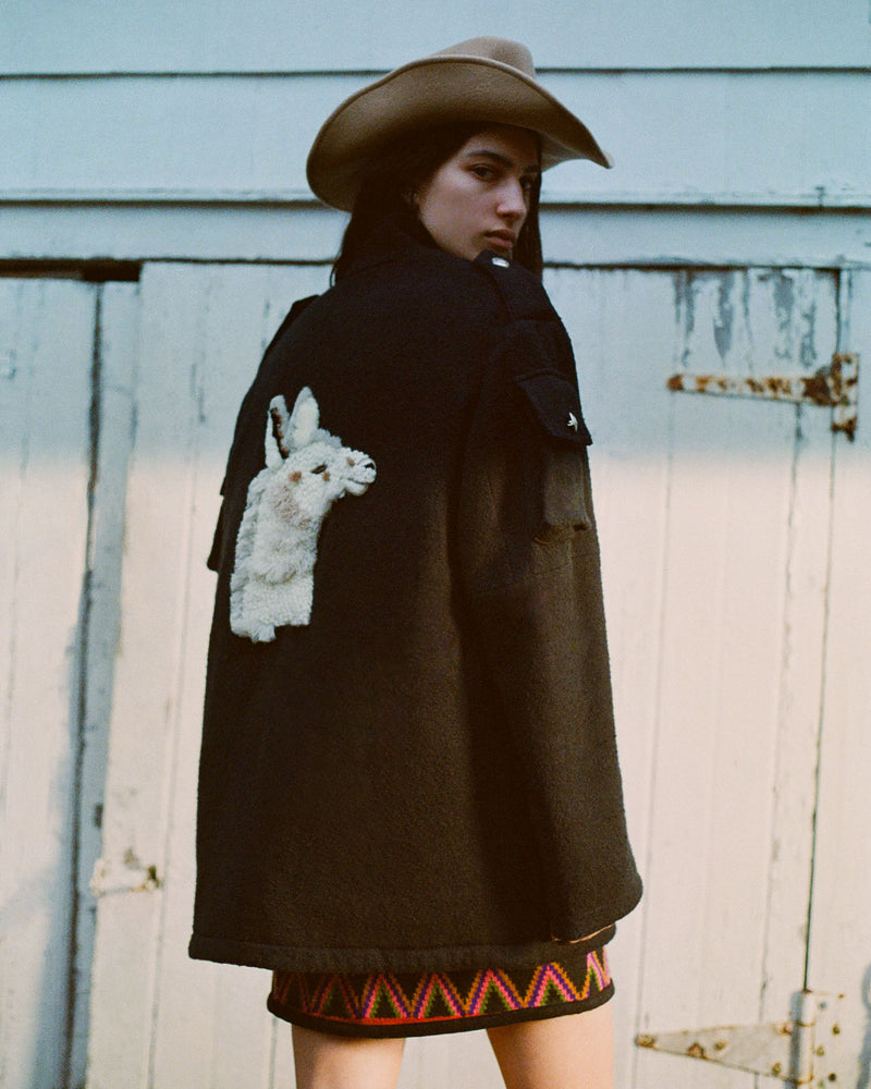 A person stands outdoors wearing a black coat with a white rabbit design on the back, a cowboy hat, and a colorful skirt. They are facing away, near a white wooden gate.