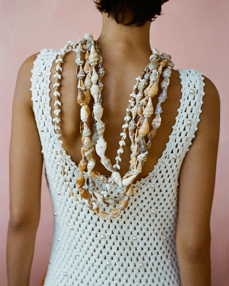 A person facing away from the camera wearing a white crocheted dress adorned with layered shell necklaces. The background is a soft pink color, highlighting the intricate details of the outfit and accessories.