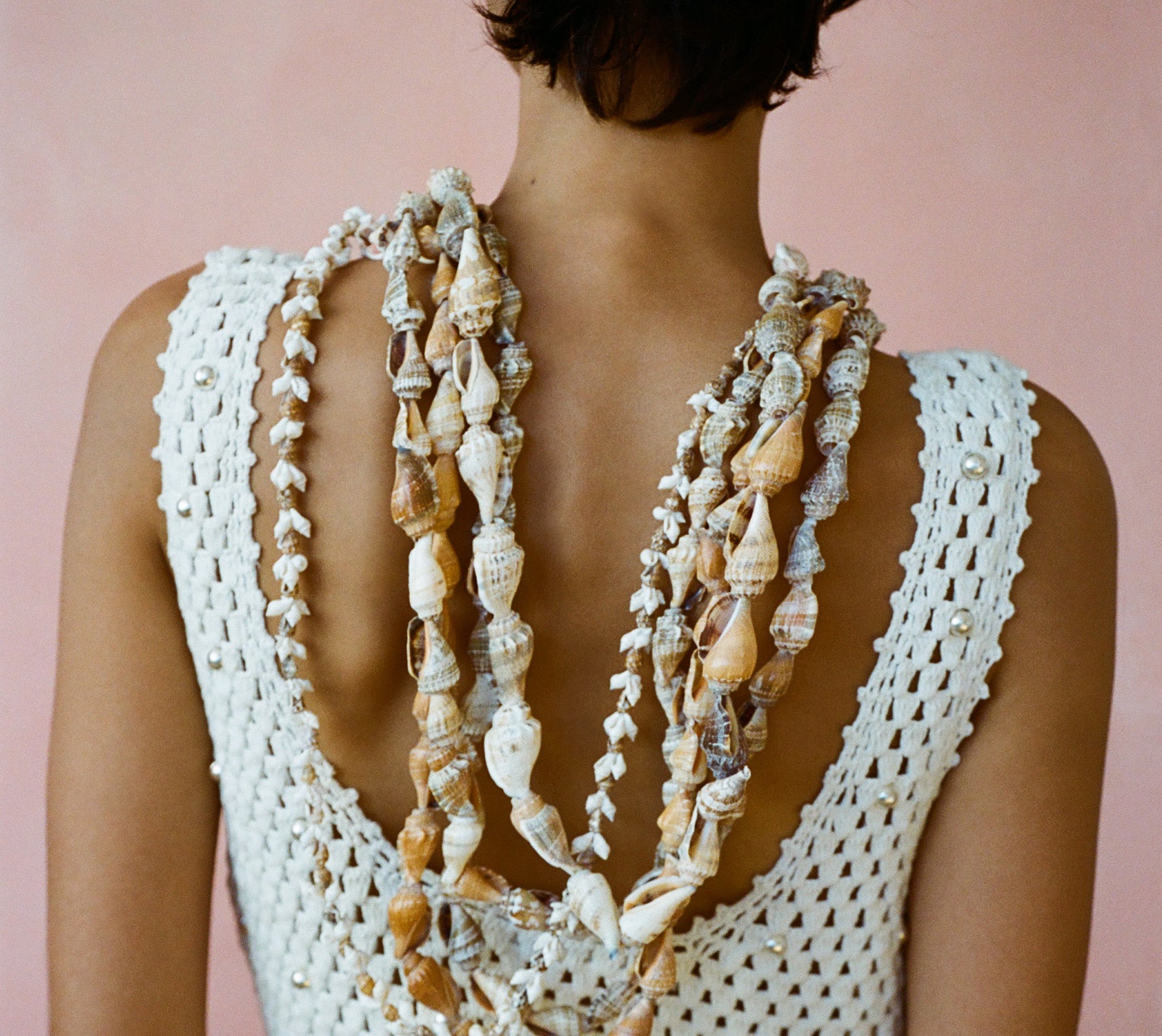 Person wearing a white crochet top with a necklace made of seashells against a pink background
