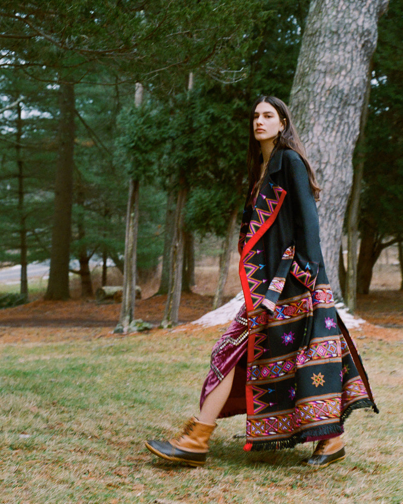 A person walks outdoors wearing a colorful, patterned long coat with geometric designs. They have long hair and are in a wooded area with trees and a soft, natural background. They're also wearing brown boots.