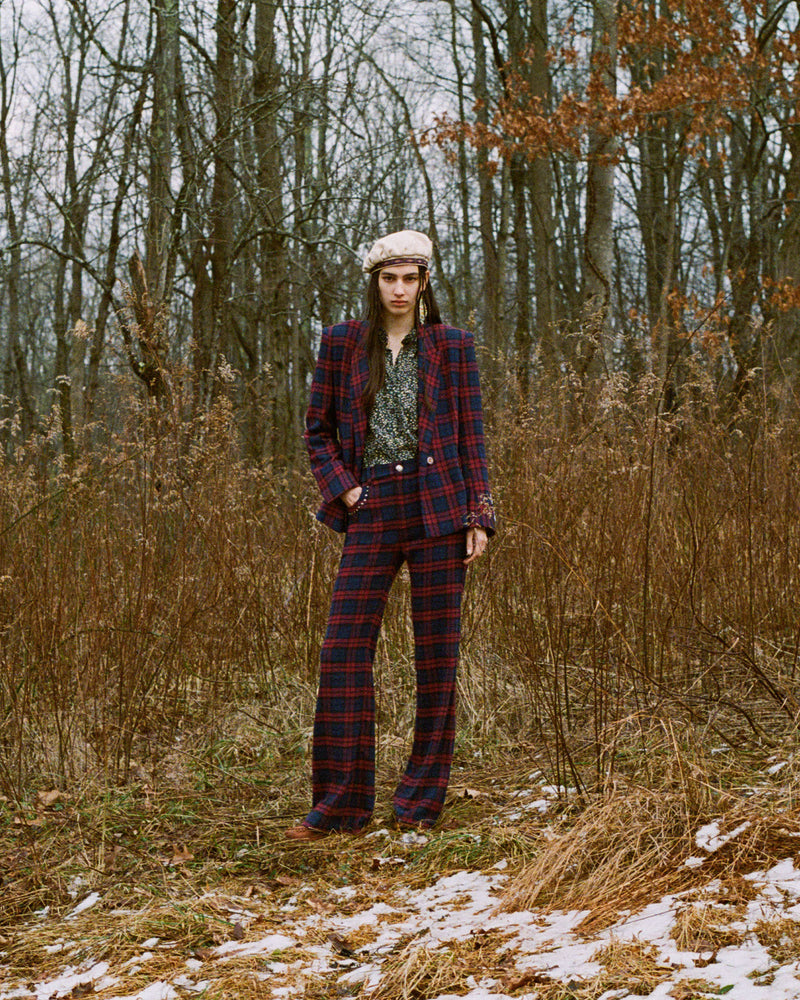 A person in a plaid suit and hat stands in a wintery forest, surrounded by sparse, leafless trees and patches of snow on the ground.