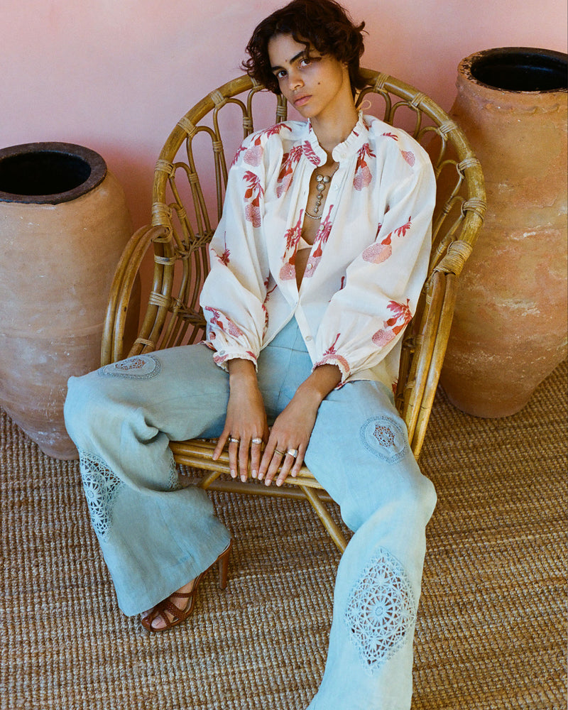 A person with curly hair sits on a wicker chair wearing a floral blouse and light blue pants with intricate lace patterns. The background features large terracotta pots and a woven rug.