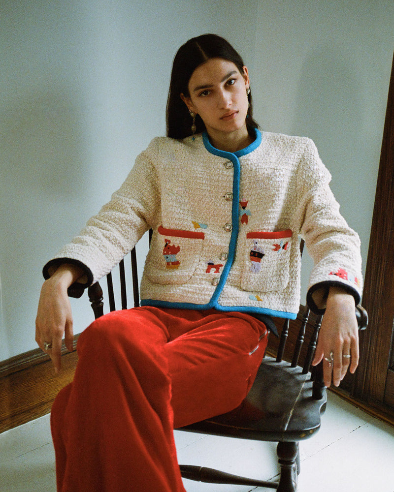 A person with long dark hair sits in a wooden chair, wearing a cream-colored textured jacket with colorful patterns and red velvet pants. The setting is a softly lit room with light walls and wooden trim.