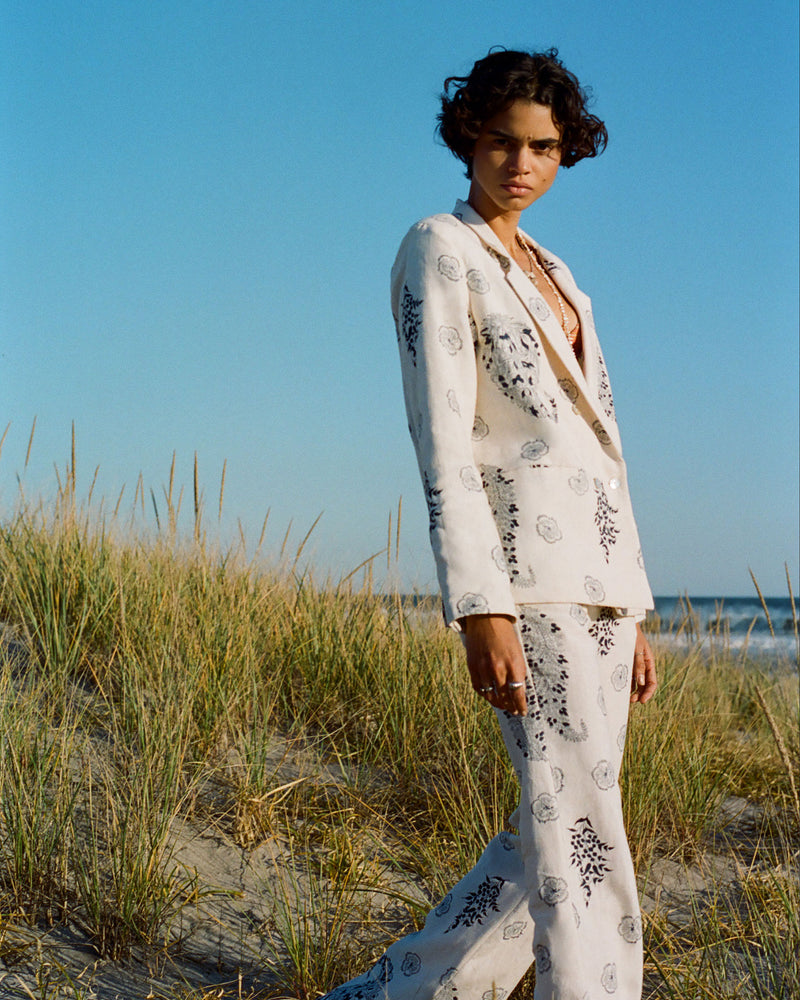 A person stands on a sandy beach surrounded by tall grasses, wearing a white suit with intricate black patterns. The background features a clear blue sky and ocean.