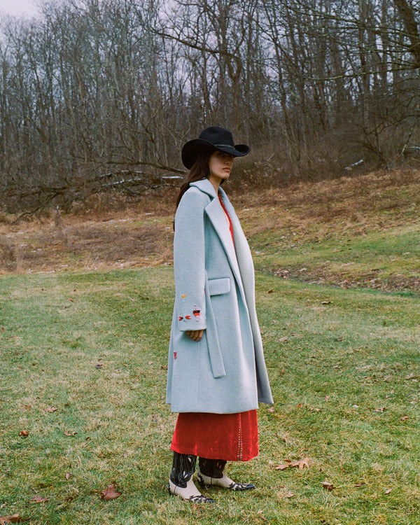 Person wearing a light blue coat and a black hat stands in a grassy field with bare trees in the background. They are also dressed in a red outfit and white boots with black details. It's an overcast day in a natural setting.