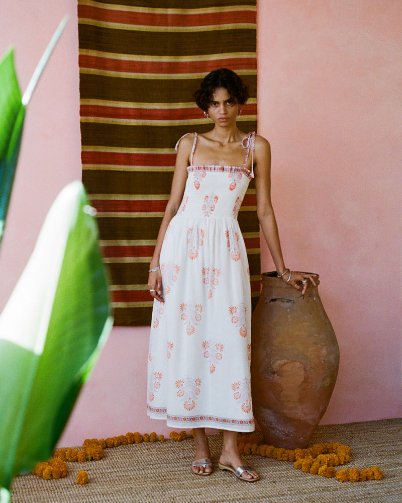 A person stands in a long, white dress with pink floral patterns, leaning on a large clay pot. The background features a textured wall and a striped hanging. Yellow flowers and a green plant are in the foreground.