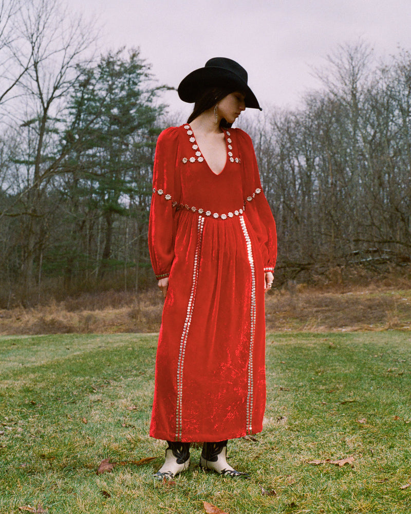 A person in a red velvet dress adorned with circular accents stands in a grassy field. They wear a wide-brimmed black hat and ankle boots. The background is a mix of bare trees and cloudy sky, suggesting a late autumn or early winter setting.