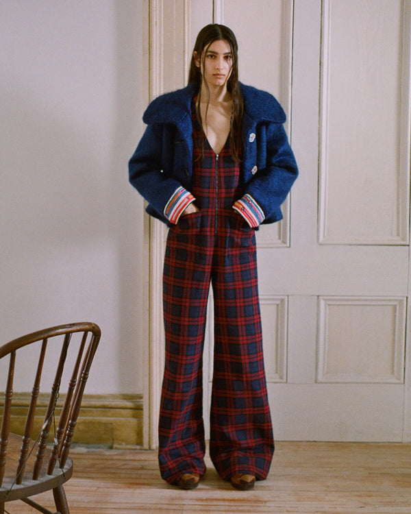 A person stands indoors wearing a blue jacket and a red and black plaid jumpsuit. Their hands are in the jacket pockets, and they are near a white door. A wooden chair is partially visible on the left.