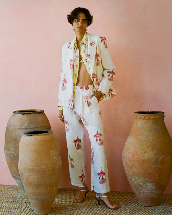 A person with short, curly hair stands against a pink backdrop wearing a white suit with red floral patterns. The suit jacket is open, revealing a patterned shirt underneath. They are flanked by three large, weathered clay pots.