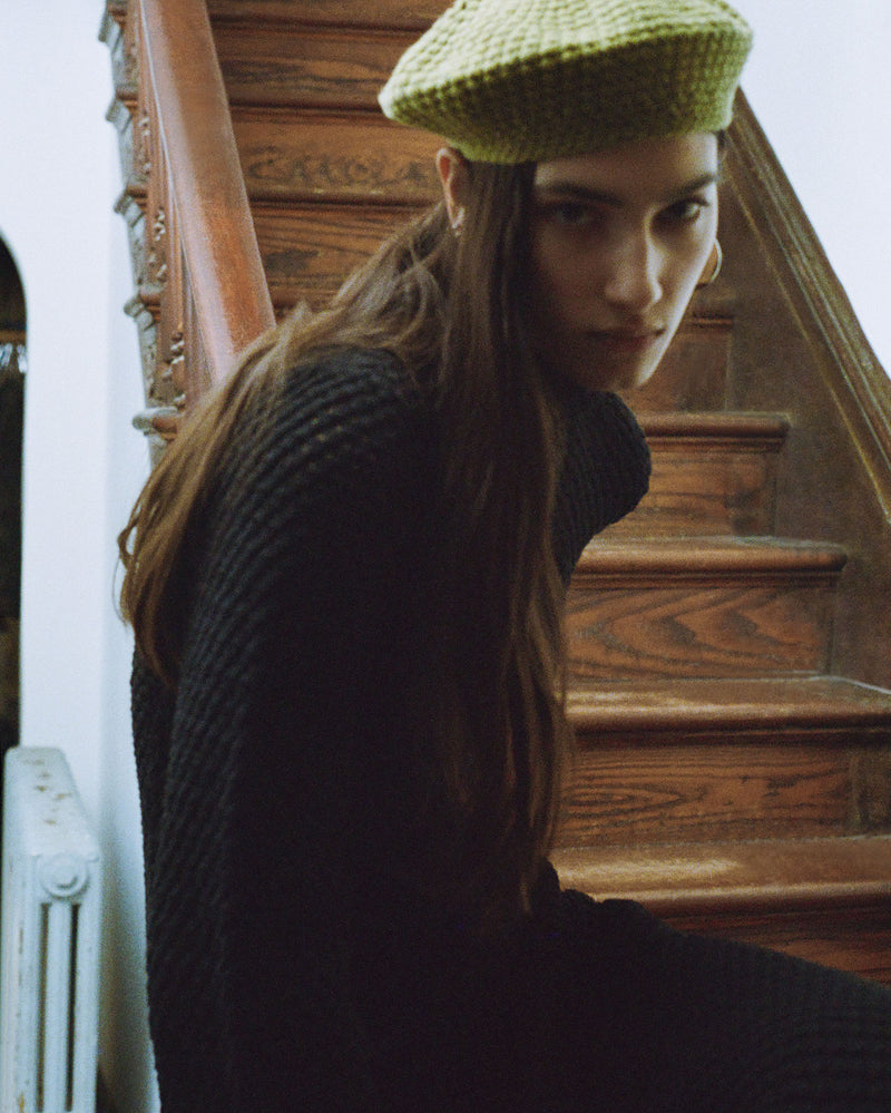 A person wearing a textured green beret and a black sweater sits on a wooden staircase, gazing towards the camera. The setting is indoors with natural light.