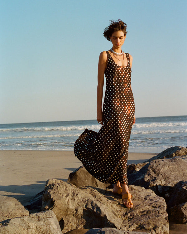 A person stands on rocks by the beach, wearing a flowing black dress with white polka dots. The ocean waves are in the background under a clear blue sky.